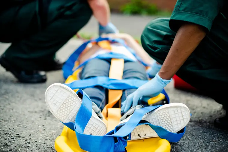 Two paramedics in green uniforms strapping a casualty onto a yellow spine board on a road.